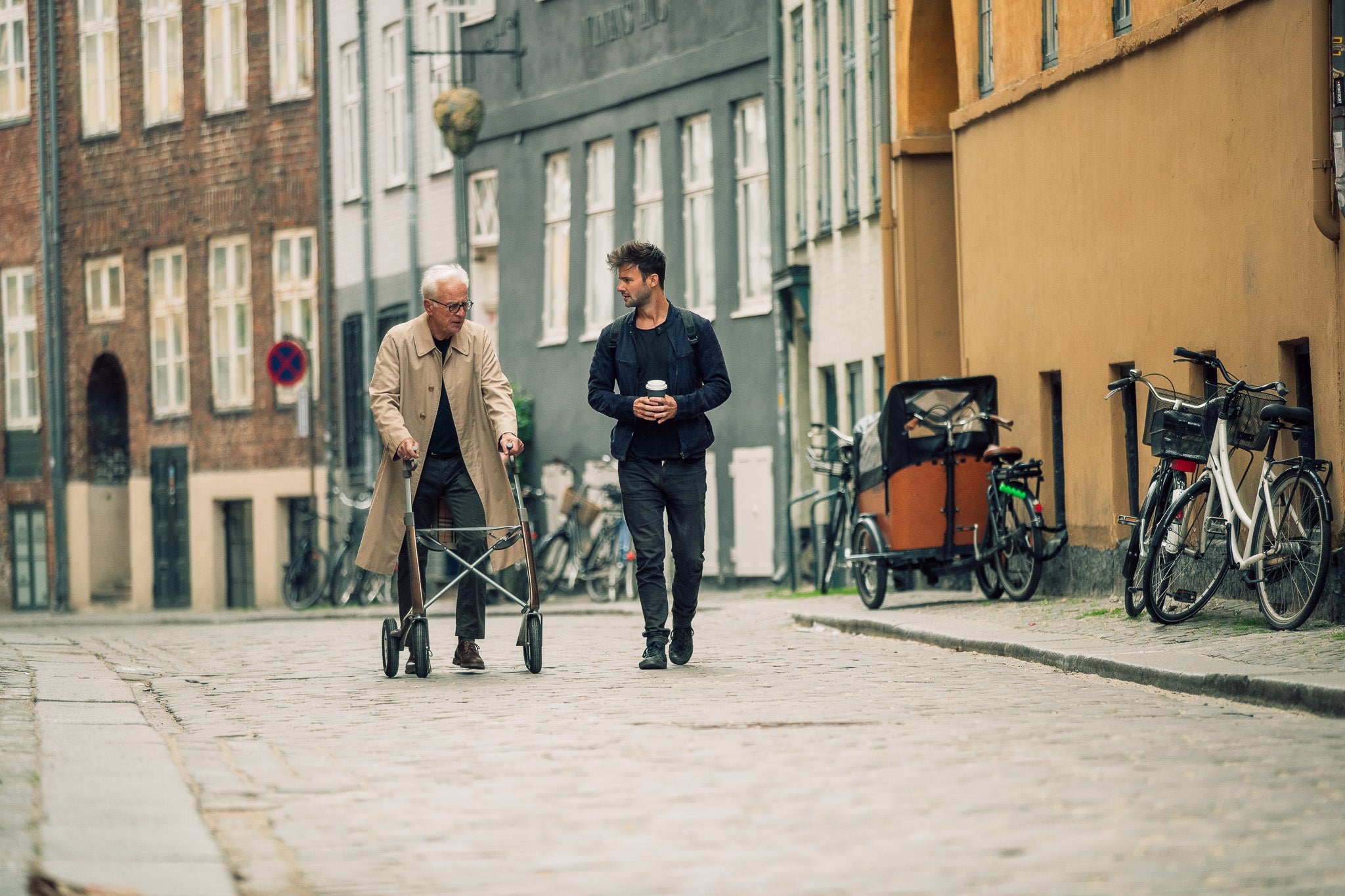 A grandfather walks with a modern lightweight walking frame along a cobble stone street, while talking to his adult grandson who walks beside him and holds a takeaway coffee. Bikes and European apartments are in the background.