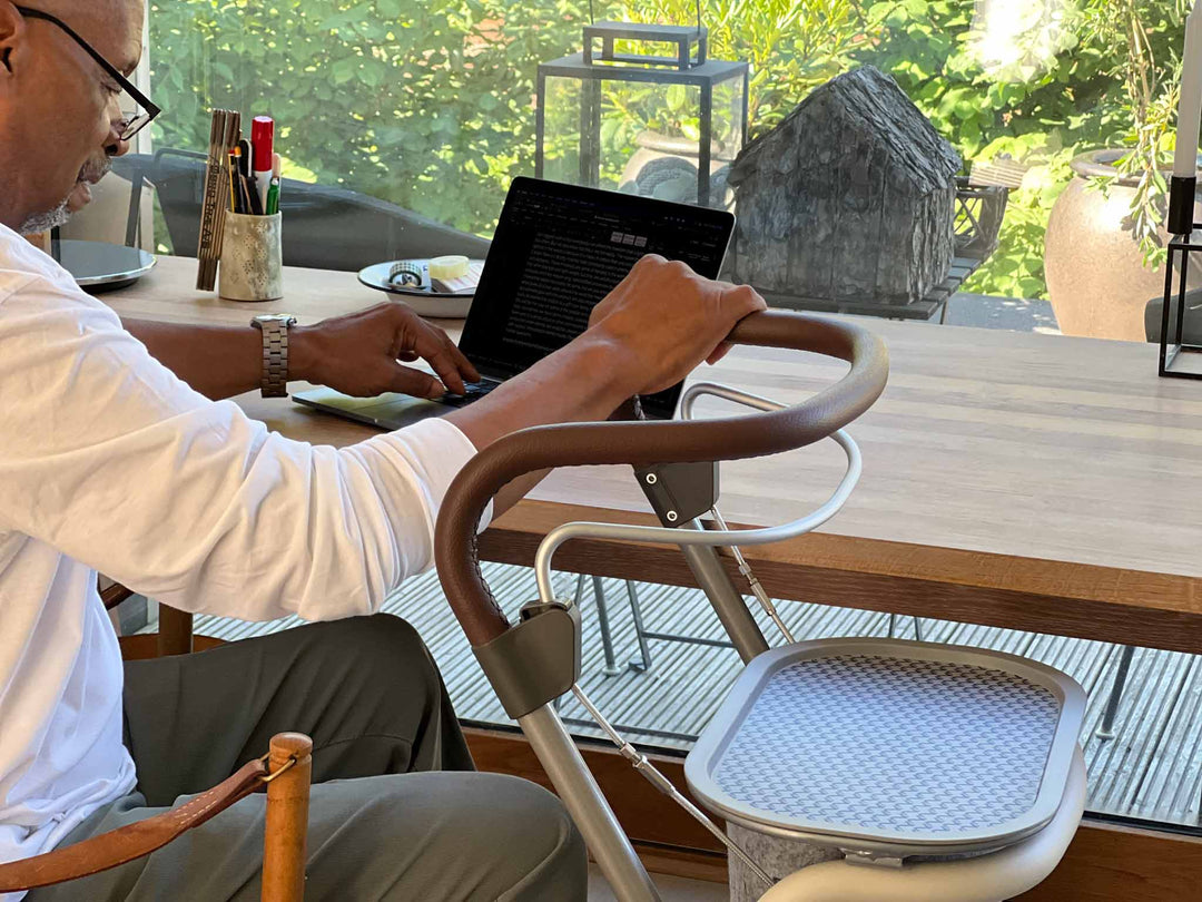 A man works on a laptop that while sitting at a desk while resting one hand on a 'byACRE Scandinavian Butler' indoor rollator