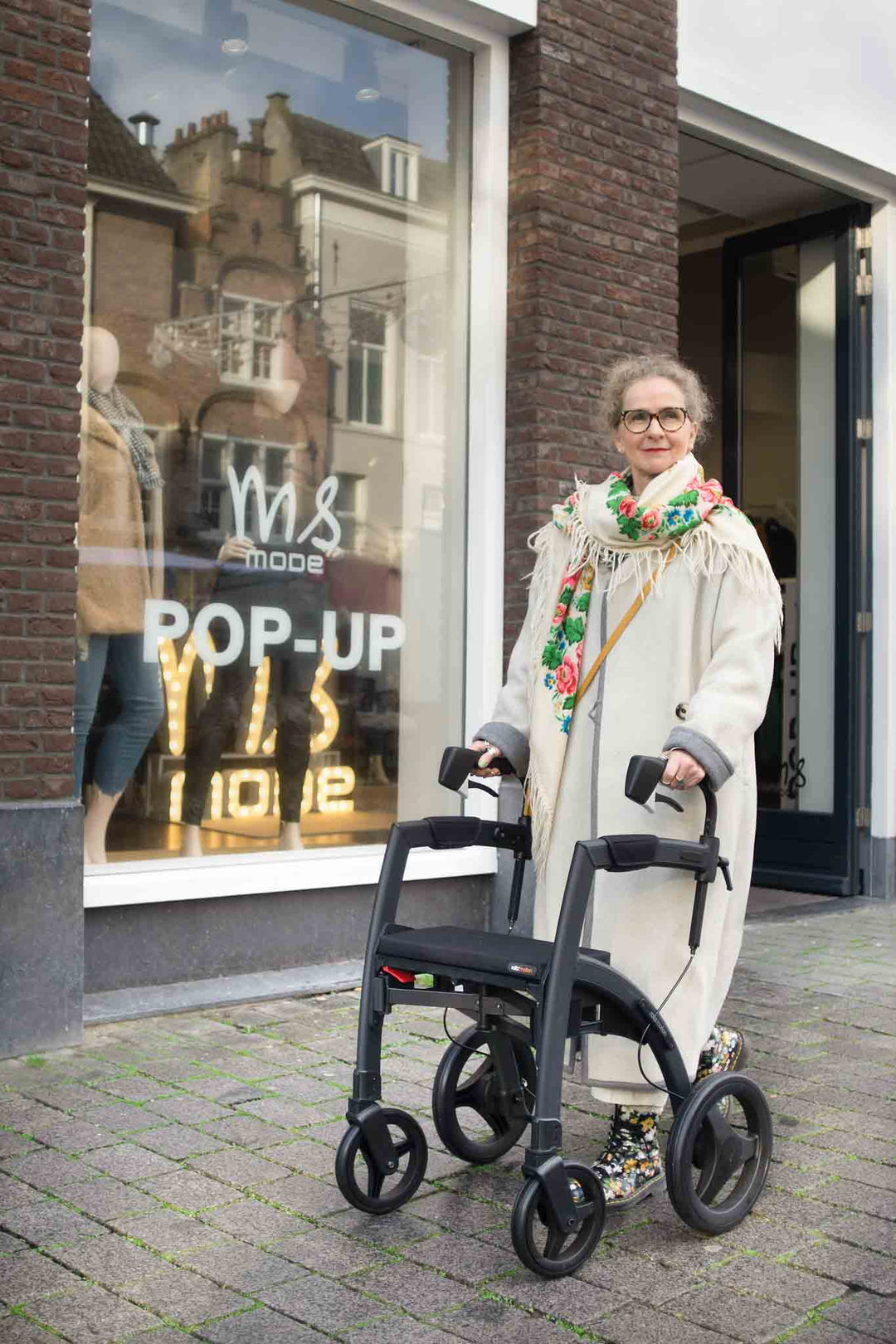 A woman stands outside a fashion shop with a 'Rollz Motion Parkinsons' walking frame