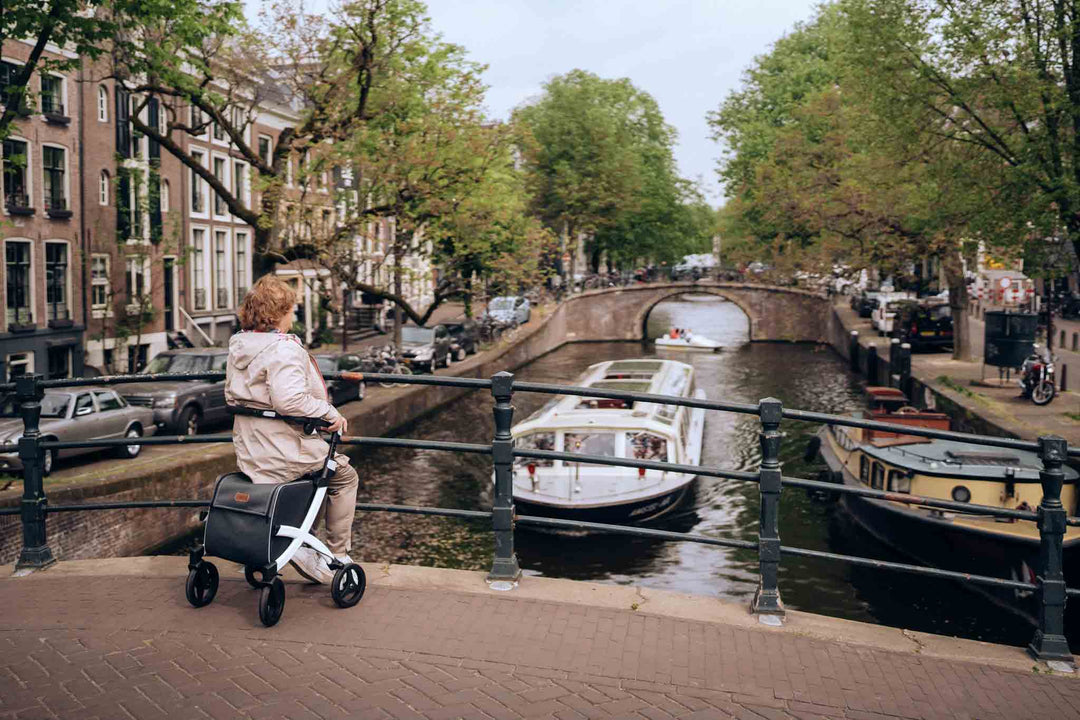A woman sits on a modern 'Rollz Flex' rollator on a bridge in Amsterdam, watching a canal boat.