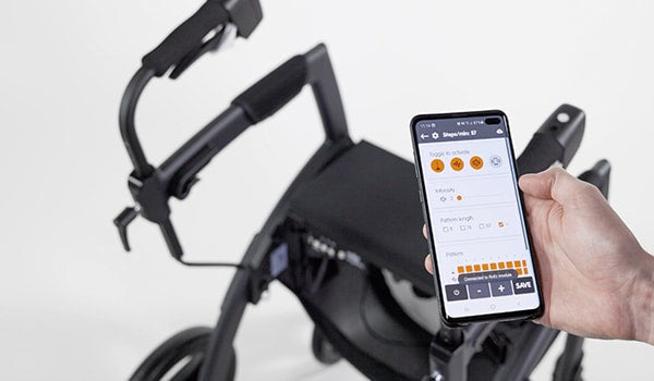 A hand holds a smart phone that displays an app that controls the cues for the Rollz Motion Rhythm, which is a walker for people with Parkinson's Disease. The walker in blurred in the background.