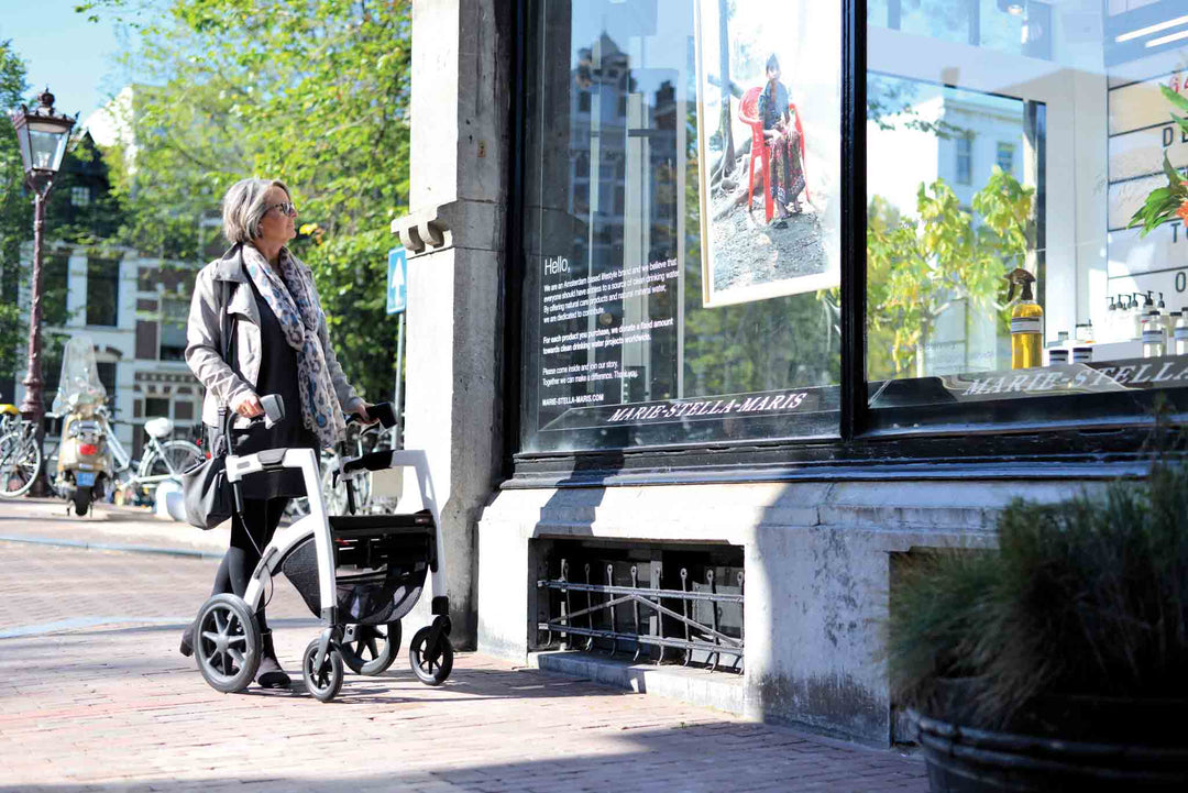 A woman walks along a footpath with a 'Rollz Motion' rollator and looks in a galley window