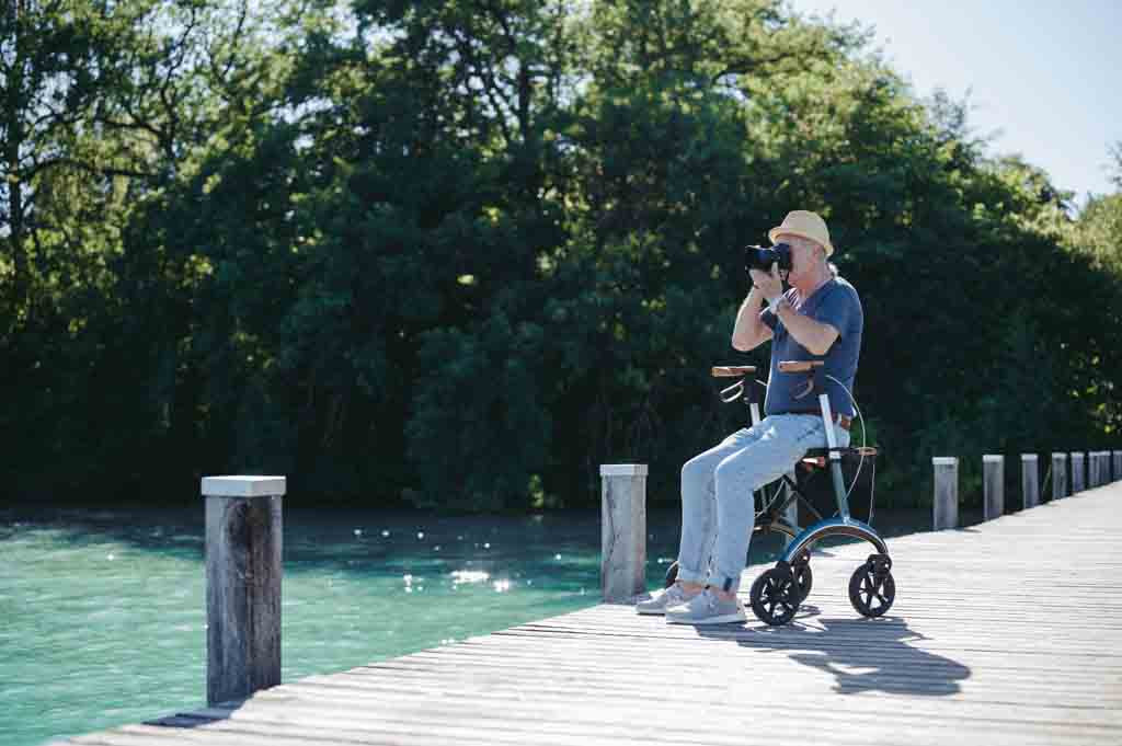 A man sits on a Saljol Carbon Fibre Walker on a jetty, taking a photograph with his camera, with trees and ocean in the background