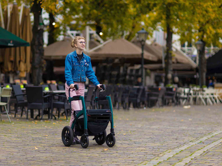 A girl walks across a brick street using a 'Rollz Motion Performance' rollator with outside seats and tables from a restaurant in the background