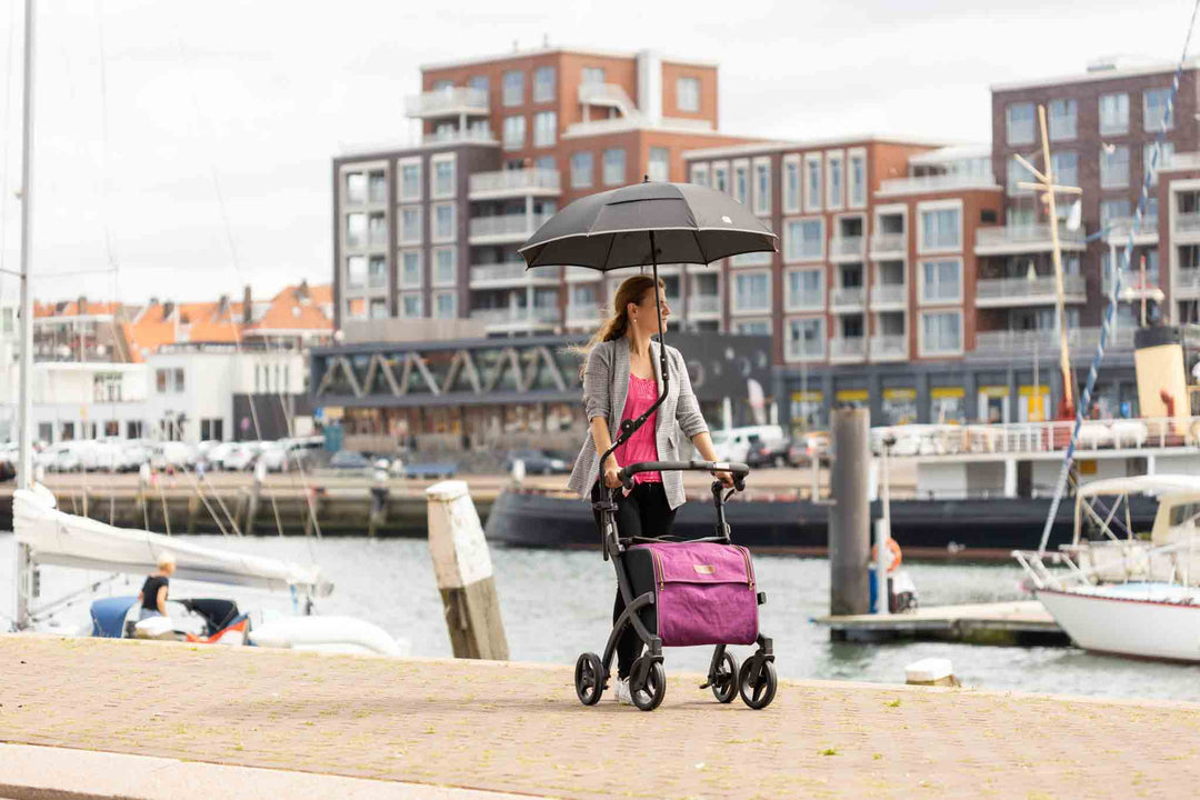 A woman walks along a footpath beside a port with the 'Rollz Flex' walking frame, with an umbrella attached, apartments and boats in the background