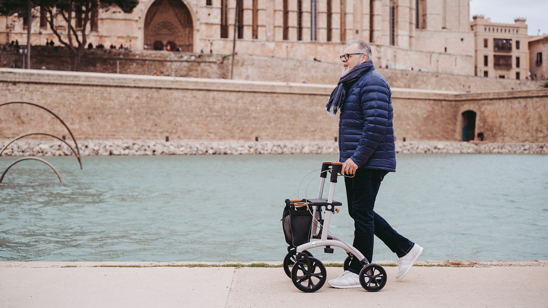 A man walks beside a river in Europe with a Saljol Allrounder rollator with a castle in the background