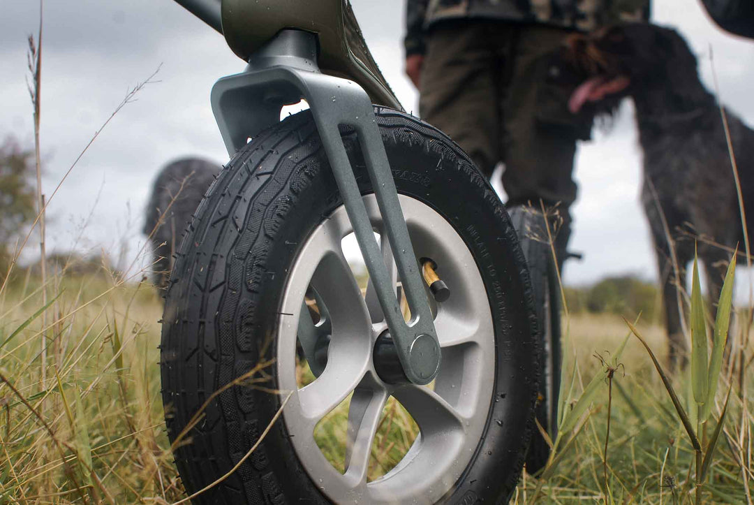 A close up of the front wheel on a 'byACRE Overland' walking frame in a grassy field with a out of focus man and dog in the background