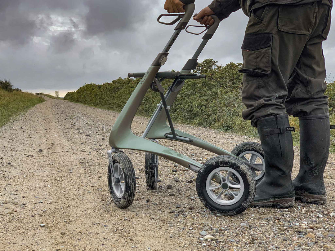 A close up of the 'byACRE Carbon Overland' walking frame, used by someone wearing gumboots on a dirt road beneath an overcast sky