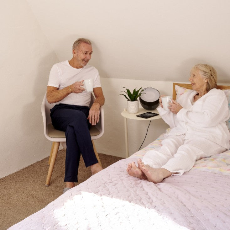An older couple in a bright bedroom. The man sits in a white chair which is also a discrete commode, holding a white mug, while the woman in a white robe relaxes on a light pink bed.