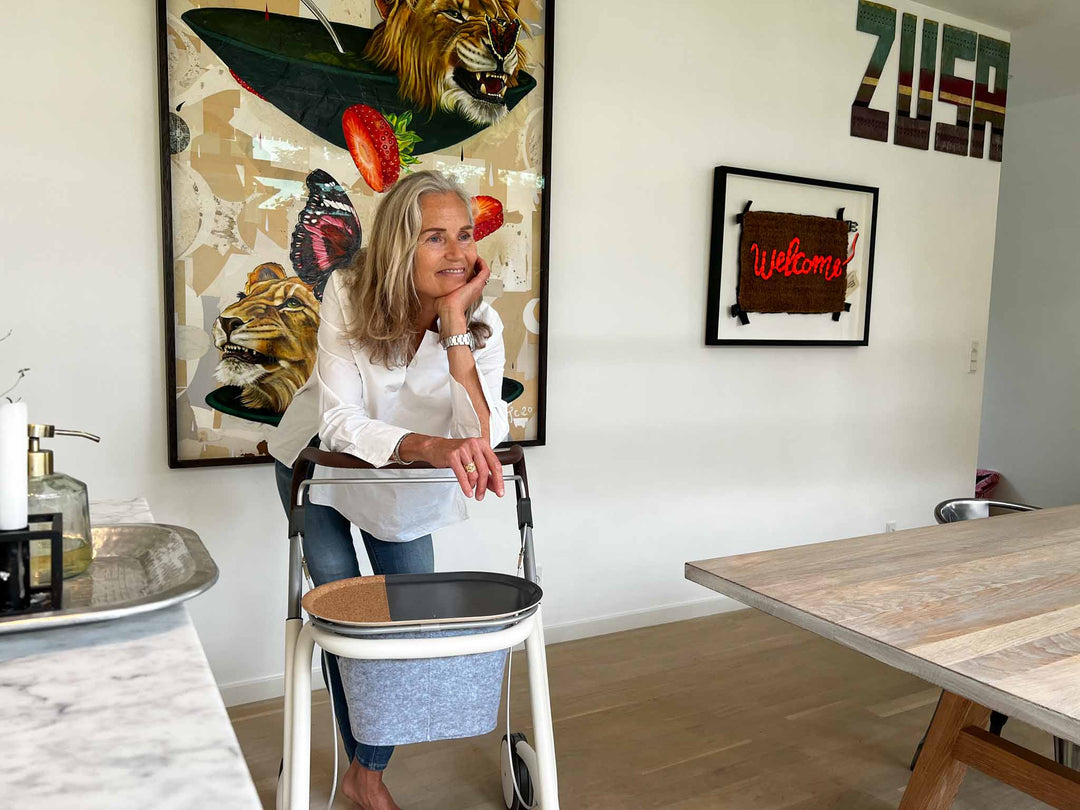 A woman relaxes with a 'byACRE Scandinavian Butler' indoor rollator in a modern kitchen with artworks on the wall behind her