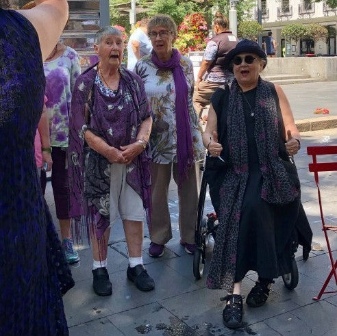 An elderly woman sits on a walking frame while singing in a town square