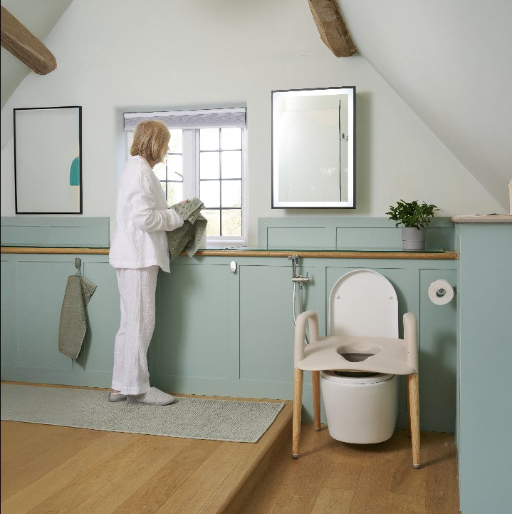 A woman in white pajamas stands by a window in a bright, modern bathroom with pale blue walls, wooden ceiling beams, a wall mirror, and a portable white raised toilet seat chair that is over a white toilet.