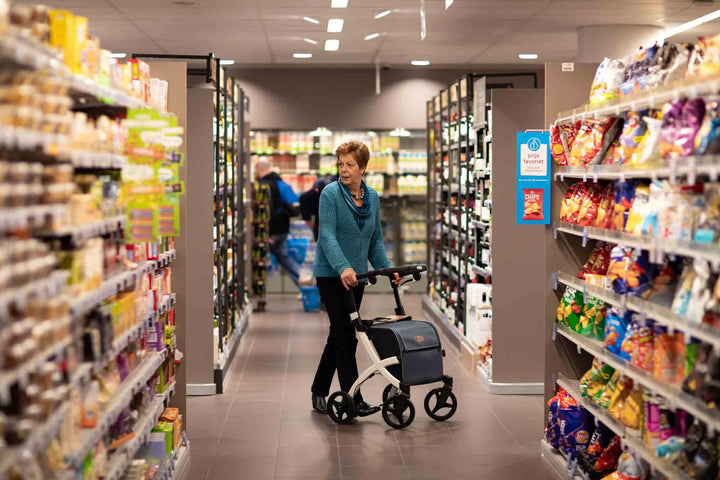 A woman walks through a supermarket using a 'Rollz Flex' rollator with shopping bag attached