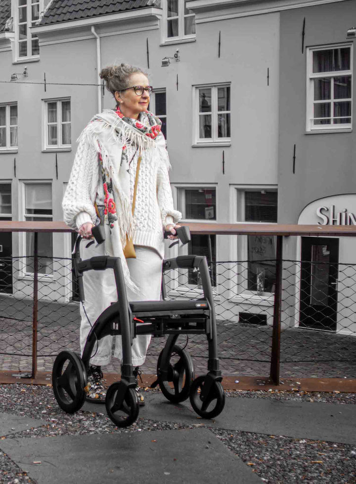 A woman walks along a footpath with the 'Rollz Motion Rhythm' walking frame with apartments in the background
