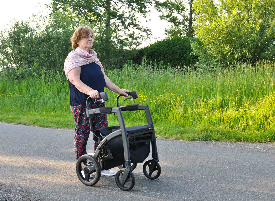A woman with Parkinson's Disease walks along a path using the 'Rollz Rhythm' walking frame with trees and grass in the background