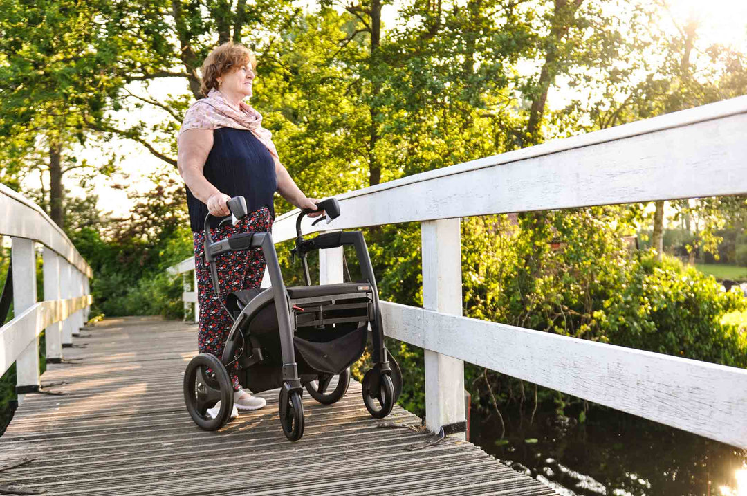 A woman who has Parkinson's Disease stands on a bridge looking at a stream while using a 'Rollz Motion' walking frame.