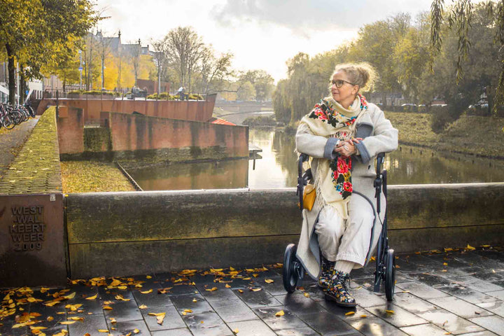 A woman sits on a walking frame on a bridge with a canal in the background