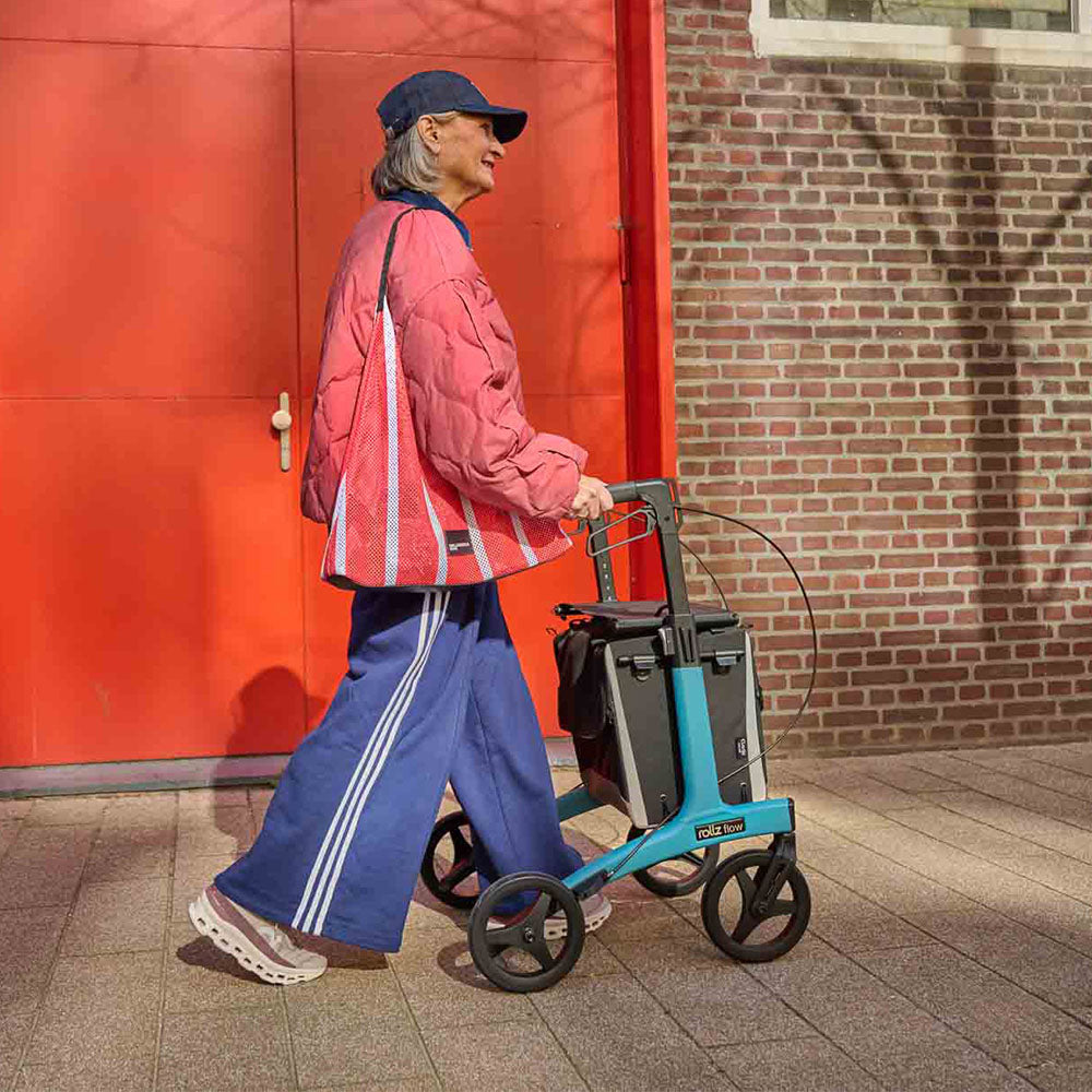 Person walking with a blue rollator on a footpath beside a red door and brick wall.