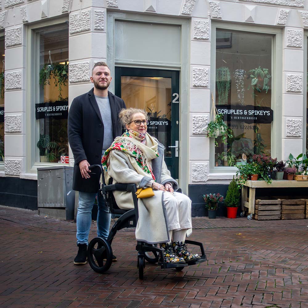 Man pushing a woman in a wheelchair on a street with a building and store front in the background.