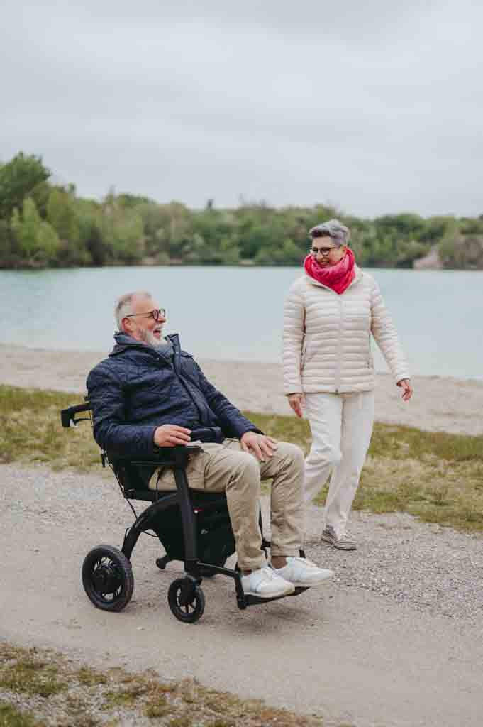 A man drive a Rollz Motion Electric wheelchair beside a lake in the company of his wife