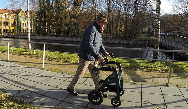 A man with Parkinson's Disease walks along a footpath with a 'Rollz Motion Rhythm' rollator , with trees and a canal in the background.