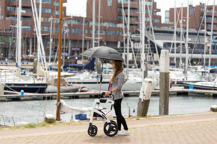 A woman walks along a dock using the 'Rollz Motion' walking frame with an umbrella attached, with boats and water in the background
