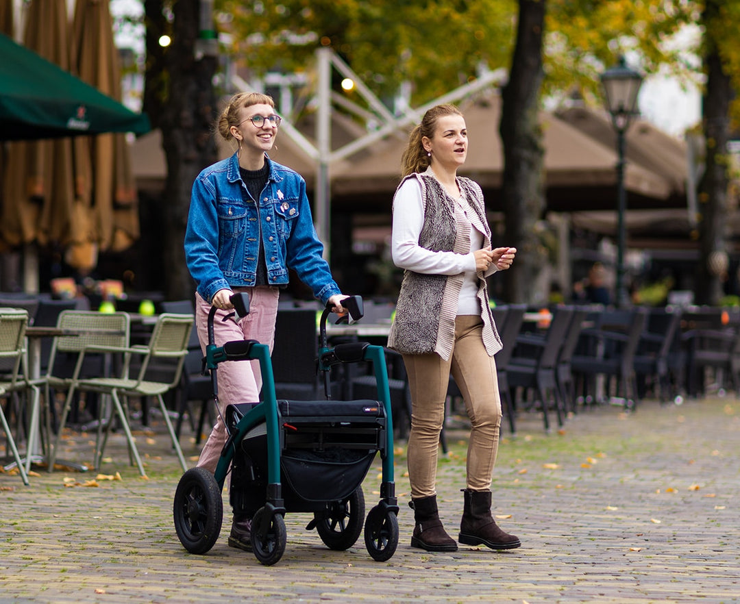 A girl walks with a 'Rollz Motion Performance' walking frame, smiling, alongside her friend, with outdoor restaurants in the background