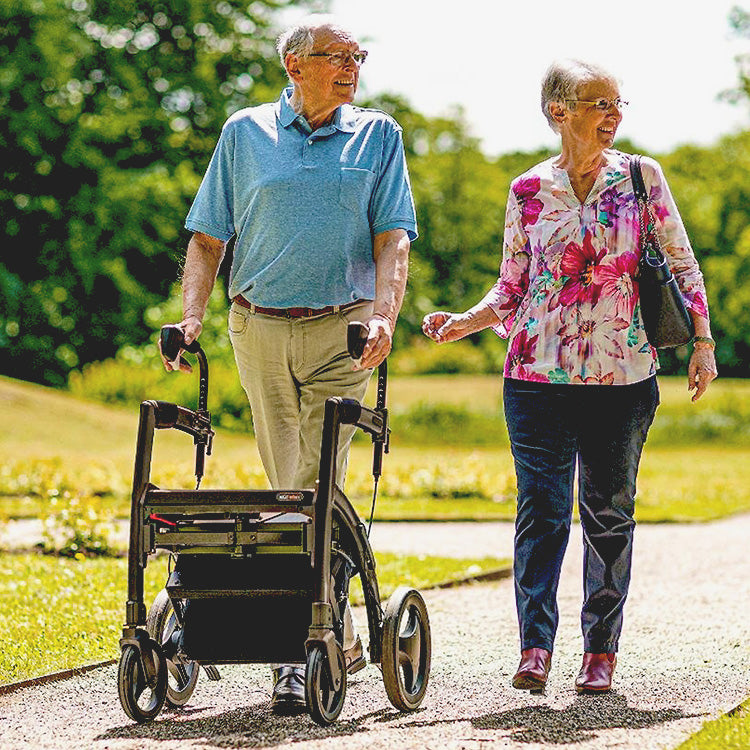 An elderly man walks with a 'Rollz Motion' walking frame that is deisgned for people with Parkinson's disease. He is accompanied by his wife, they are both smiling, strolling in the park with trees in the background.