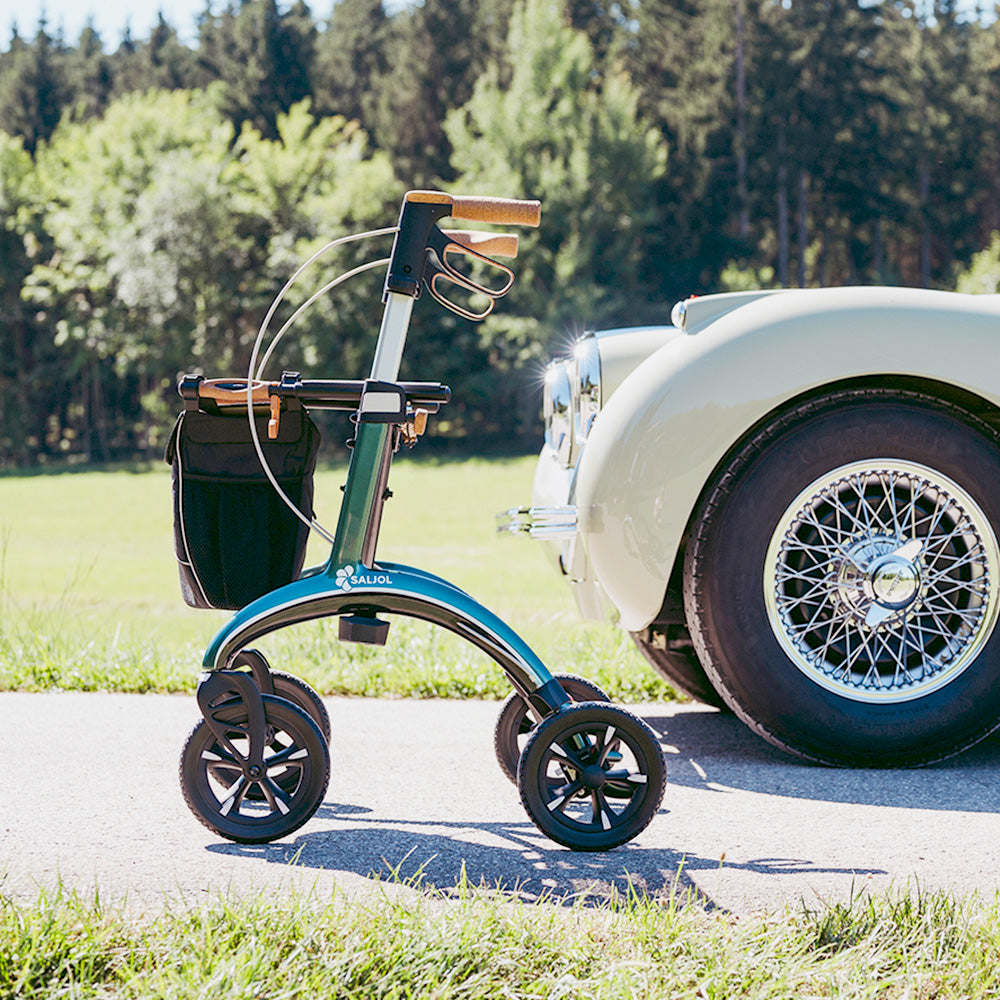 A green carbon fibre Saljol rollator is on a driveway with a classic car and trees in the background