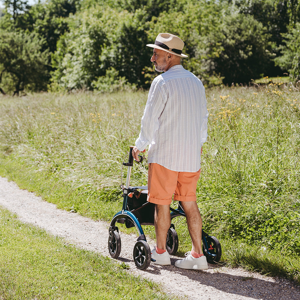 A man walks down a pebbly path surrounded by a field, while using a Saljol Carbon Fibre Rollator