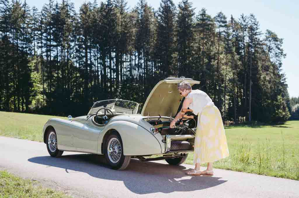 A woman puts a Saljol rollator in the boot of a classic car on a driveway with trees in the background