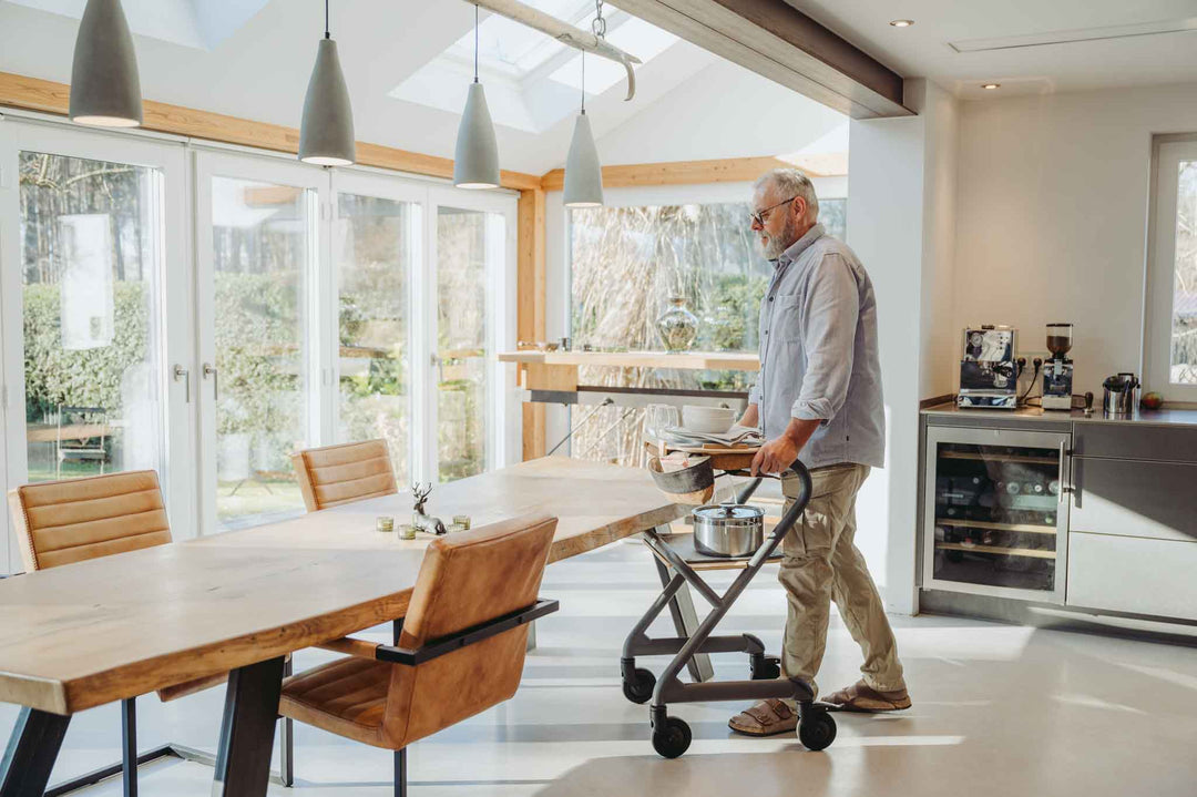 A man transports food and plates to a dining table in a modern house while using a Saljol indoor walker for stability.