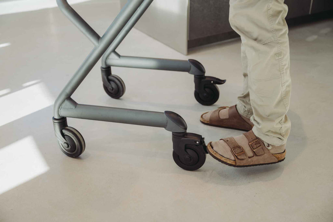 Person wearing sandals adjusts the rear wheel of an indoor mobility aid, on a light-colored floor.