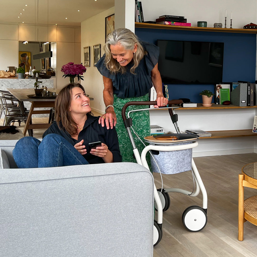 A woman laughs with her daughter in a loungeroom, standing beside her indoor walking frame made by 'byACRE'