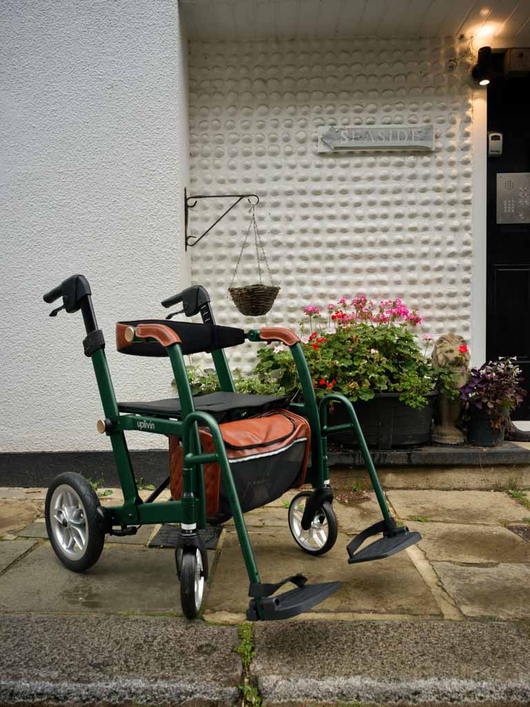Green mobility walker wheelchair with seat, on a concrete surface, with a white wall and plants in the background.