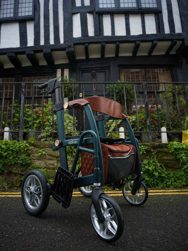 Green and brown 2-in-1 walker wheelchair with a bag in front of an old English building.