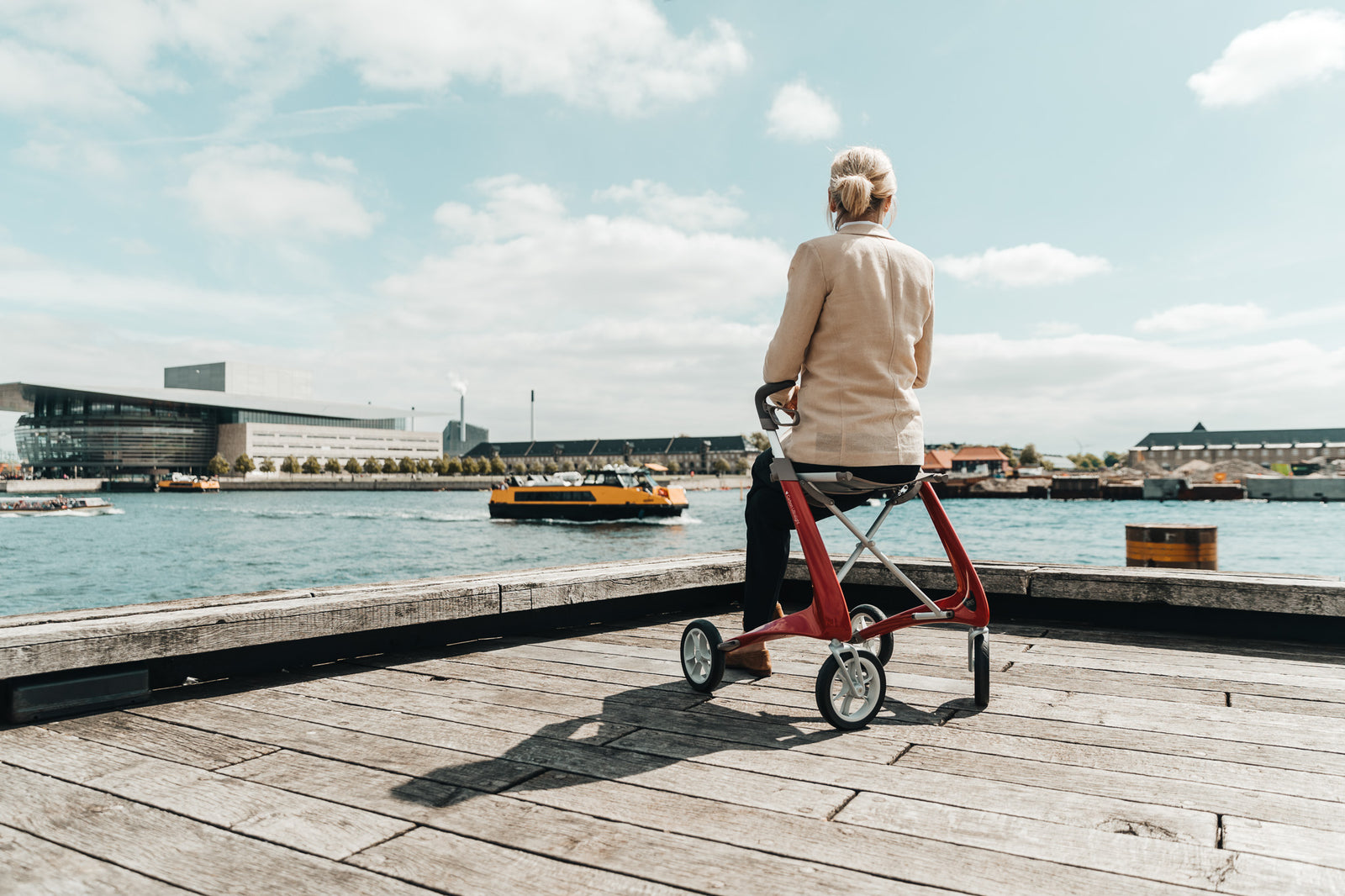 A woman sits on a modern light-weight walking frame, made by byACRE. She is on a timber wharf looking out at a harbour where a yellow ferry passes in front of the Copenhagen Opera House, on a sunny day with blue sky