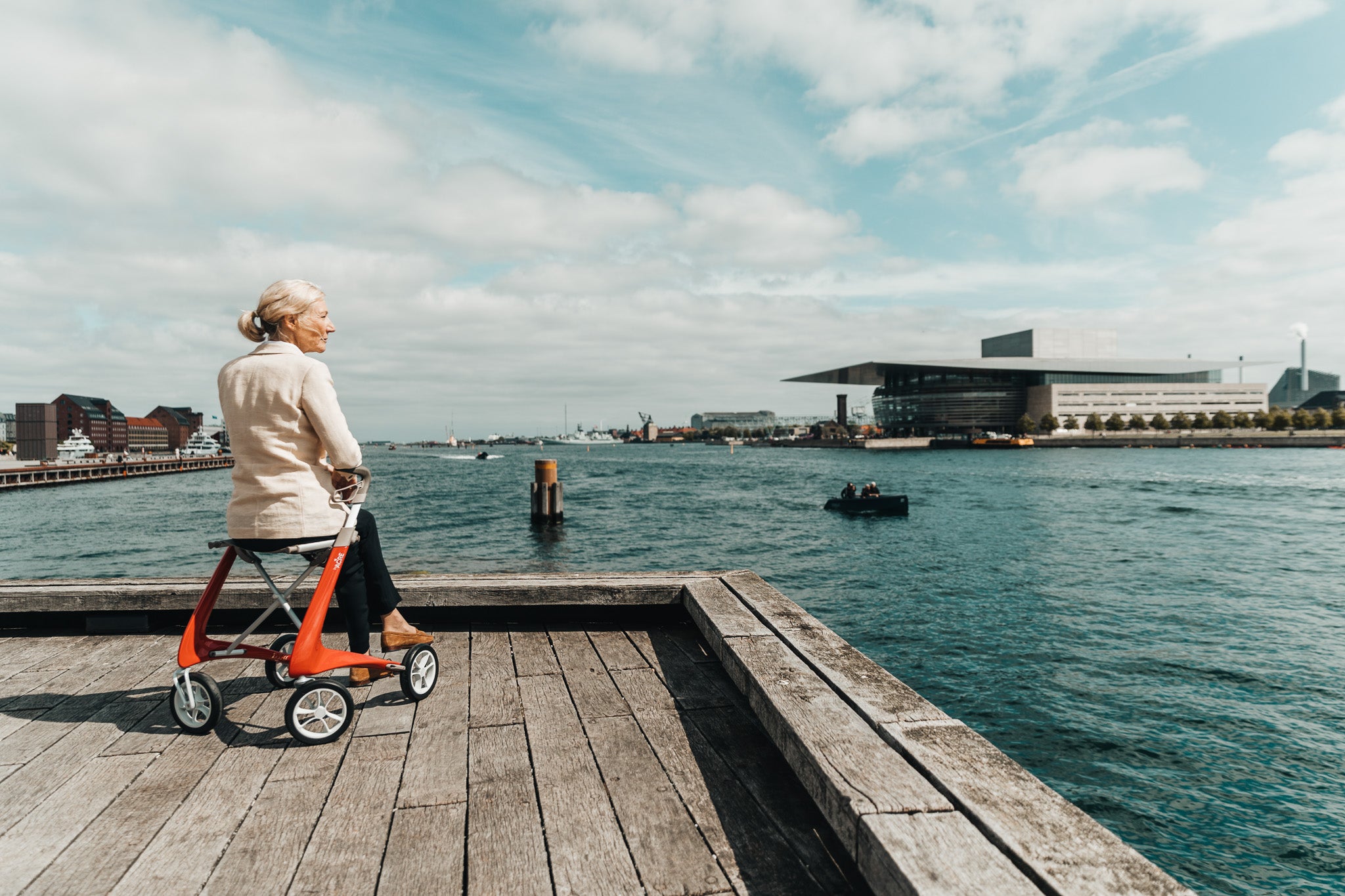 A woman sits on a red 'byACRE Carbon Ultralight' walking frame, on a jetty, with the Copenhagen Opera House in the background