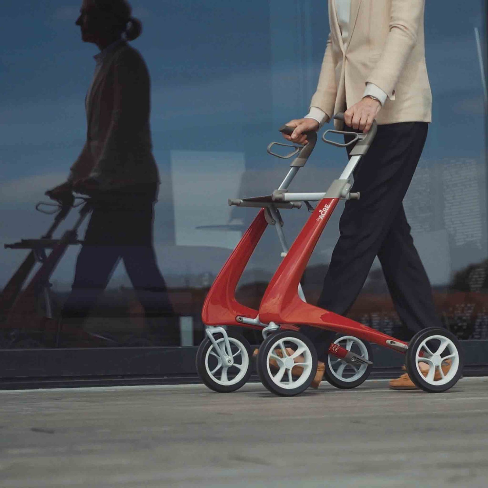 A close up of a red 'byACRE Ultraight' walking frame being used by a woman walking past a window