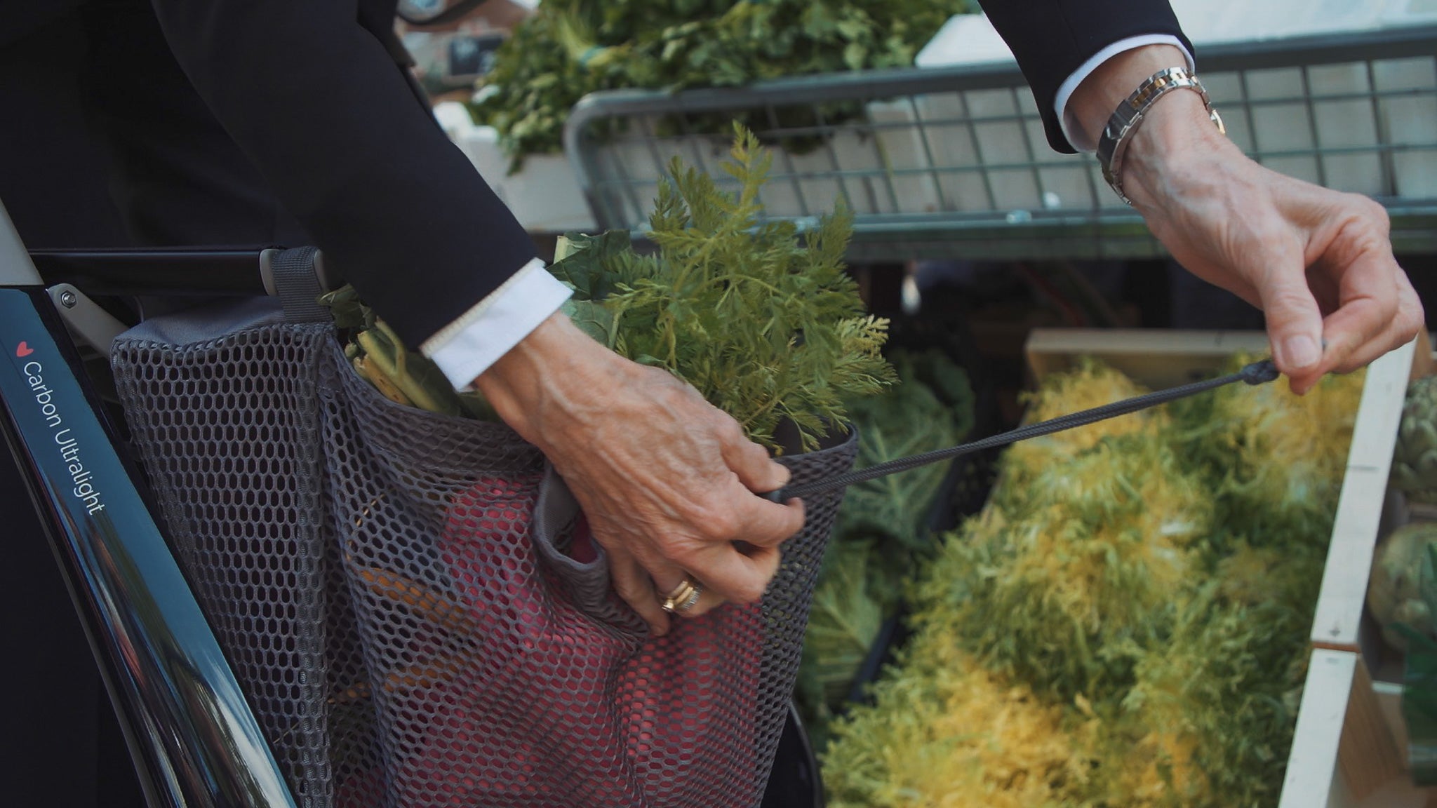 A close up of a mesh shopping bag attached to a walking frame. The bag is carrying carrots and is being closed by a woman's hands.