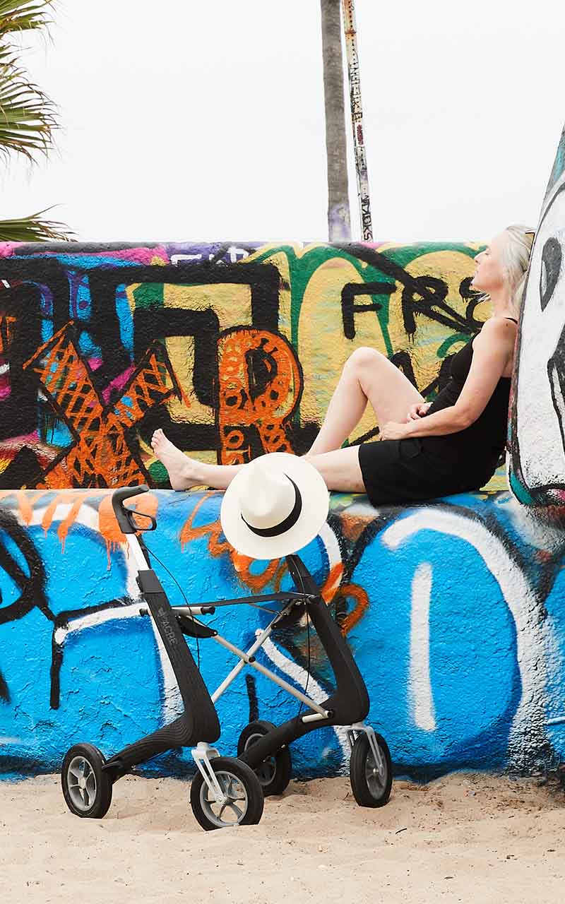 A woman rests on top of a concreate wall that is covered in graffiti, at the beach, with a byACRE walking frame beside her.