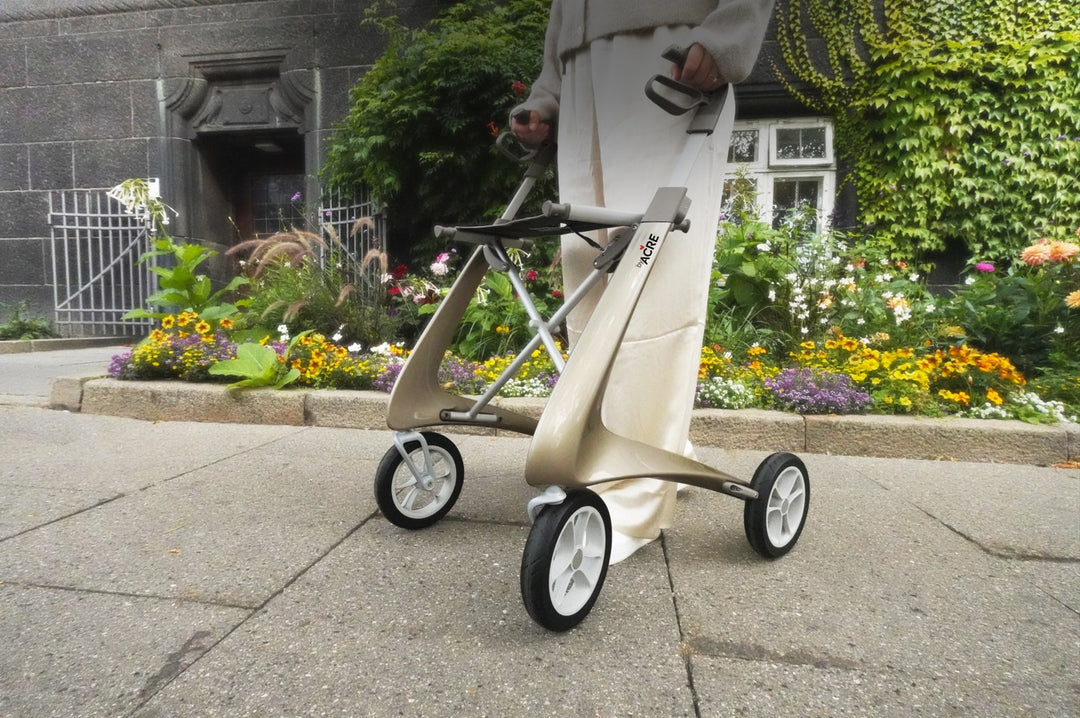 Person pushing a futuristic, compact walker on a sidewalk with flowers and a building in the background.