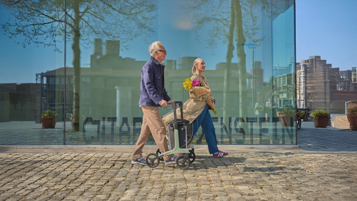 Two people walking with a walker in front of a glass building.
