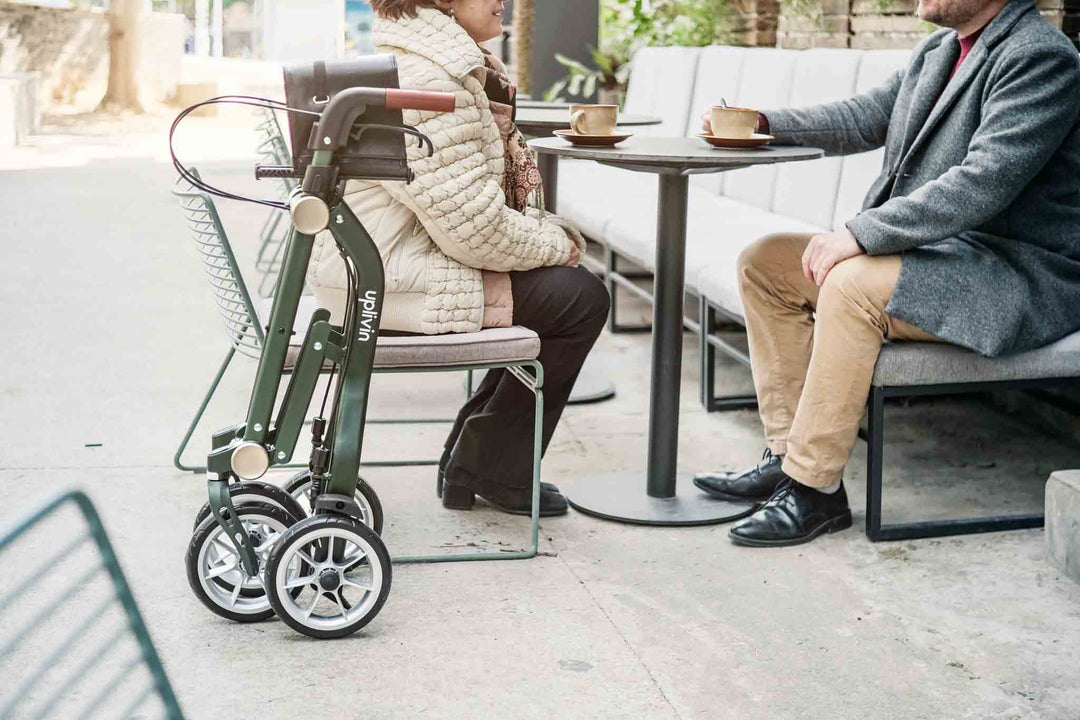 A green walking-frame is folded and stands beside a table where a lady and man sit enjoying coffee