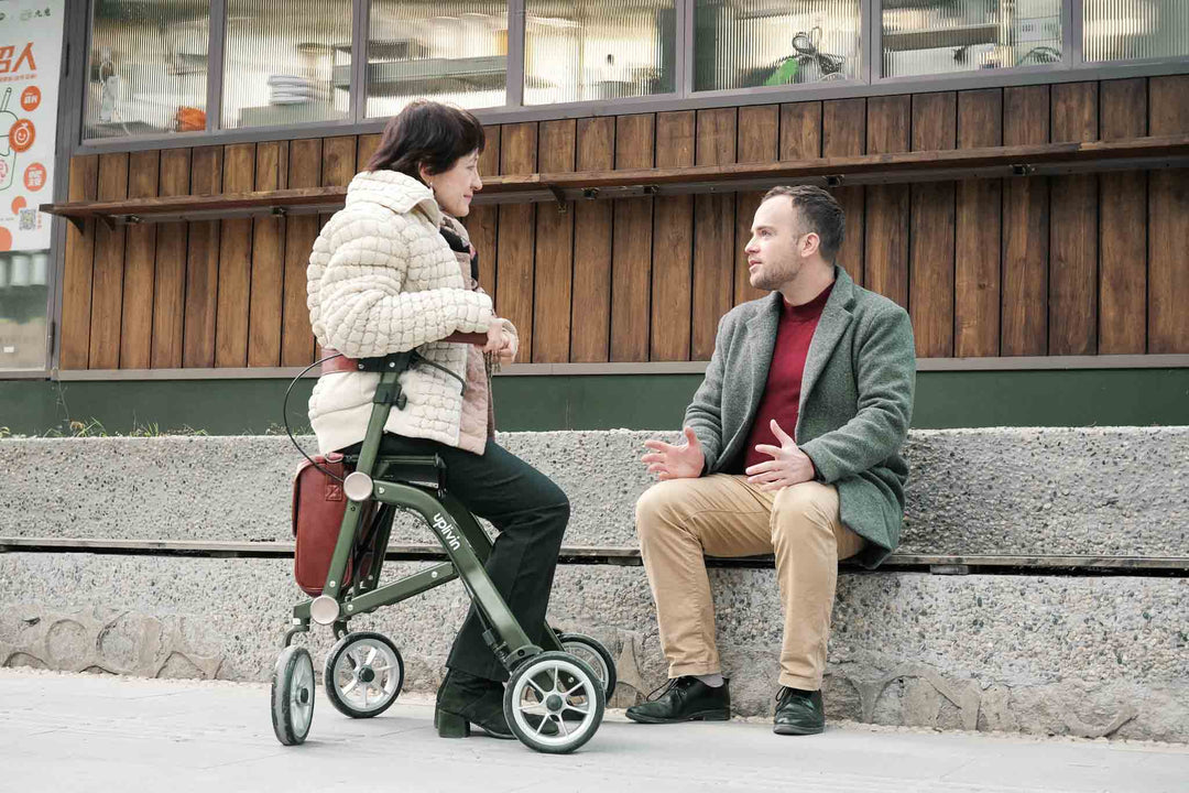 A woman sits on a green 'Uplivin Trive' walking frame and talks to a man who sits on a large step in front of a building