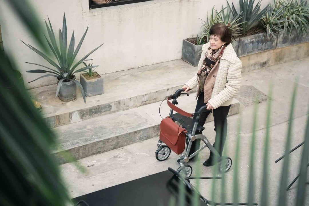 A woman walks along a path using an 'Uplivin Trive' walker rollator with a modern bag attached. 