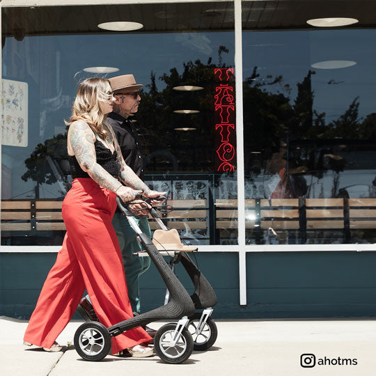 A woman walks along a footpath with a fasionable looking walker rollator with a tattoo parlor in the background